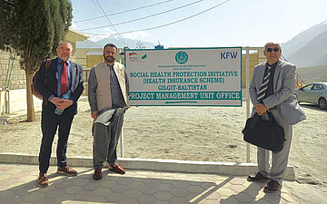 Foto von Ahmed Ishtiaq (m.), Leiter der Sehat-Sahulat-Krankenversicherung in Gilgit-Baltistan, mit Poul Thim (l.) und Dr. Brig Habib Ur Rehman von der Beraterfirma m4h vor einem Schild der KfW-Bank an einem staubigen Parkplatz
