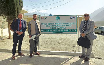 Pakistan Foto von Ahmed Ishtiaq (m.), Leiter der Sehat-Sahulat-Krankenversicherung in Gilgit-Baltistan, mit Poul Thim (l.) und Dr. Brig Habib Ur Rehman von der Beraterfirma m4h vor einem Schild der KfW-Bank an einem staubigen Parkplatz