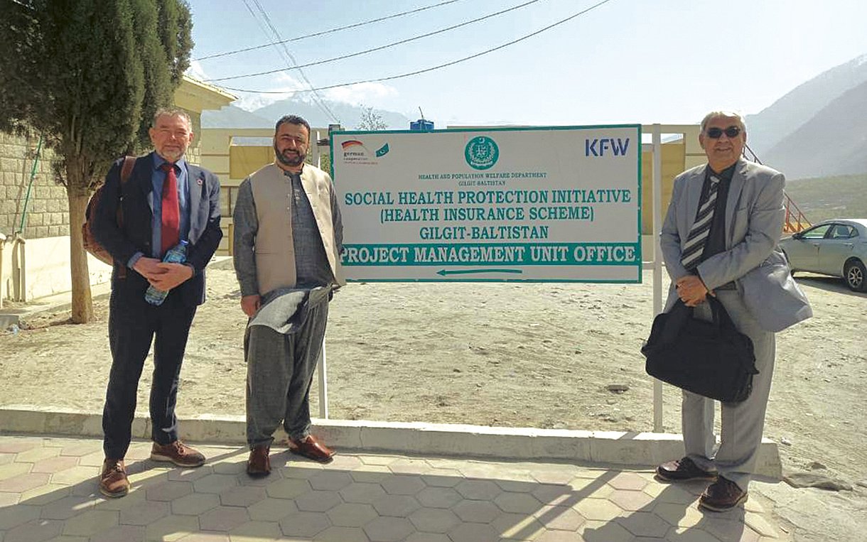 Foto von Ahmed Ishtiaq (m.), Leiter der Sehat-Sahulat-Krankenversicherung in Gilgit-Baltistan, mit Poul Thim (l.) und Dr. Brig Habib Ur Rehman von der Beraterfirma m4h vor einem Schild der KfW-Bank an einem staubigen Parkplatz