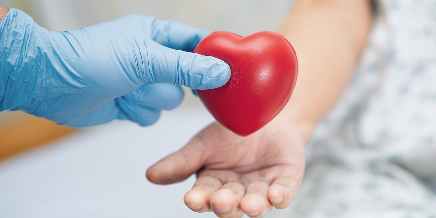 Foto: Eine Hand mit einem blauen Einmalhandschuh übergibt ein rotes Gummiherz an eine offen gehaltene Hand.