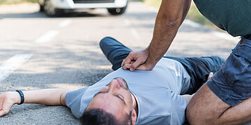 Ein Mann liegt auf dem R&uuml;cken auf der Stra&szlig;e. neben ihm kniet ein Mann und f&uuml;hrt eine Herzdruckmassage durch.