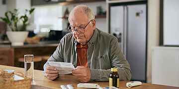Ein &auml;lterer Herr sitzt am Tisch mit Tabletten vor sich und liest eine Packungsbeilage.
