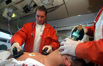 Das Bild zeigt zwei Rettungssanit&auml;ter im Rettungswagen bei der Wiederbelebung eines Patienten mit Defibrilator und Sauerstoffsack