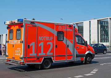 Ein Rettungswagen in roter Leuchtfarbe und mit gro&szlig;er aufgedruckter Notrufnummer 112 an den Seiten, f&auml;hrt im Einsatz eine Stra&szlig;e entlang.