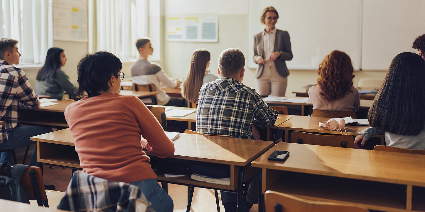 Foto: Schülerinnen und Schüler sitzen an Tischen in einem Klassenraum, vor ihnen steht eine Frau, die Lehrerin.