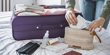 Auf einem Bett liegt ein gepackter Koffer. Daneben steht eine Frau, die gerade Medikamente in eine kleine Tasche packt.