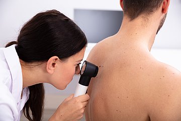 Ärztin führt Hautkrebsscreening am Patienten durch Eine Ärztin sucht mit einem Vergrößerungsglas die Haut eines Patienten nach Auffälligkeiten ab
