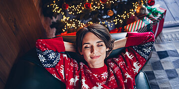Foto: Eine Frau im Weihnachtspullover liegt entspannt mit geschlossenen Augen auf der Couch. 