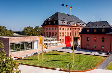 Das Foto zeigt zwei rot-braune Geb&auml;ude mit schwarzem Schifer bedecktem Haus. Links ist ein modernes Funktionsgeb&auml;ude mit gro&szlig;er Fensterfront zu sehen.