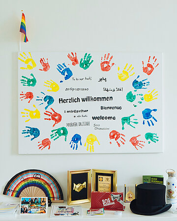 Ein Bild mit "Herzlich Willkommen" in verschiedenen Sprachen und vielen verschiedenfarbigen Handabdr&uuml;cken h&auml;ngt an der Wand. Auf dem Tisch davor stehen verschiedene Dinge, zum Beispiel ein F&auml;cher oder ein Zylinder der Schornsteinfegerinnung.