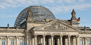 Das Foto zeigt den Blick auf das den oberen Teil des S&auml;ulenportals des Reichtagsgeb&auml;udes in Berlin mit der Aufschrift "Dem deutschen Volke". Dahinter erhebt sich die gl&auml;serne Reichtagskuppel 