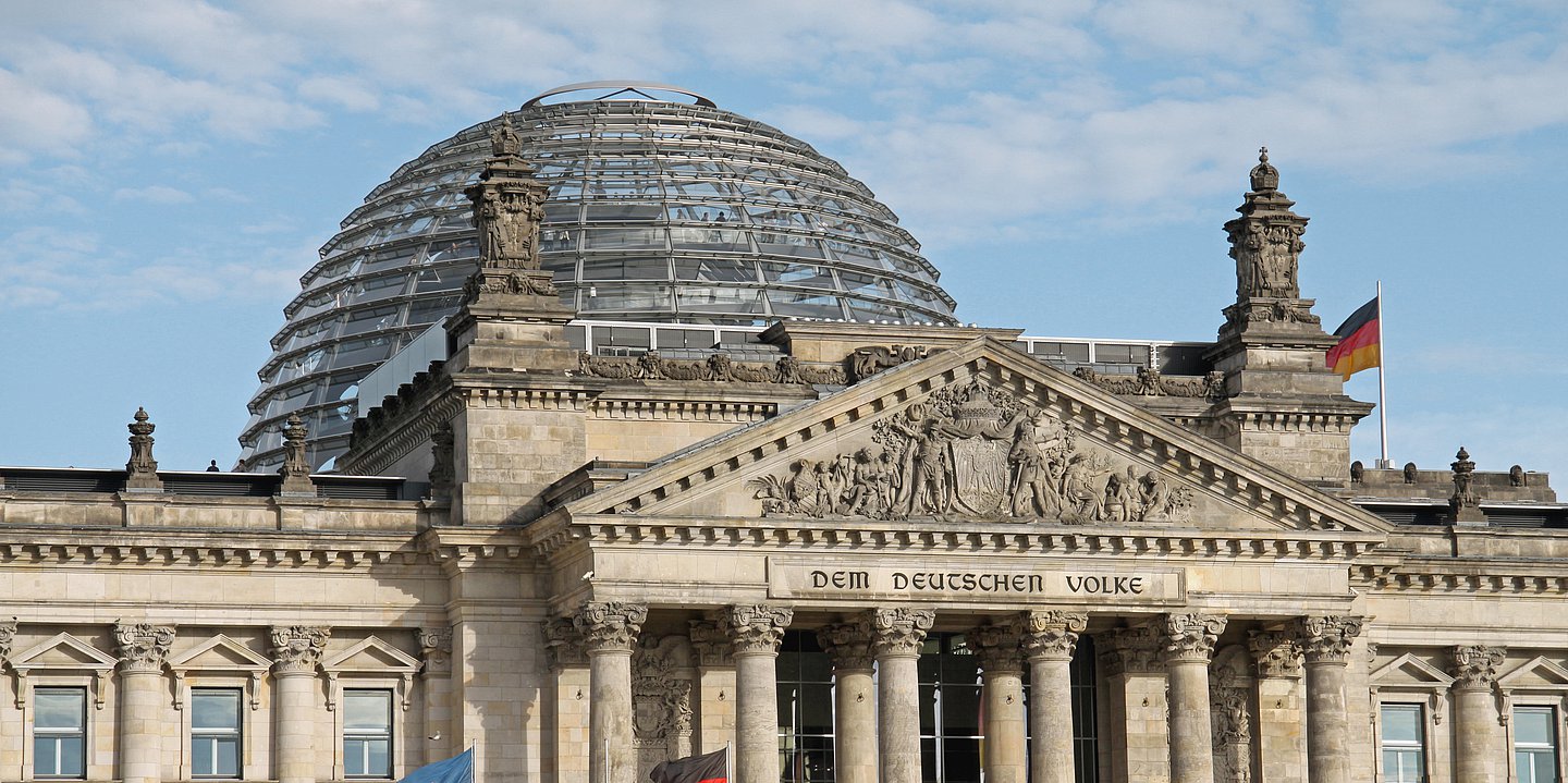 Das Foto zeigt den Blick auf das den oberen Teil des Säulenportals des Reichtagsgebäudes in Berlin mit der Aufschrift "Dem deutschen Volke". Dahinter erhebt sich die gläserne Reichtagskuppel 