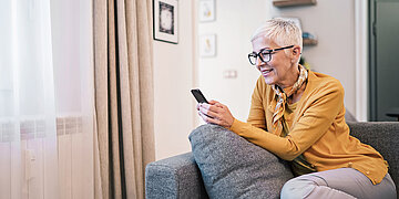 Eine Frau mit kurzen, hellgrauen Haaren in gelber Strickjacke sitzt auf einer grauen Couch mit F&uuml;&szlig;en oben und schaut in ihr Handy. Sie l&auml;chelt.