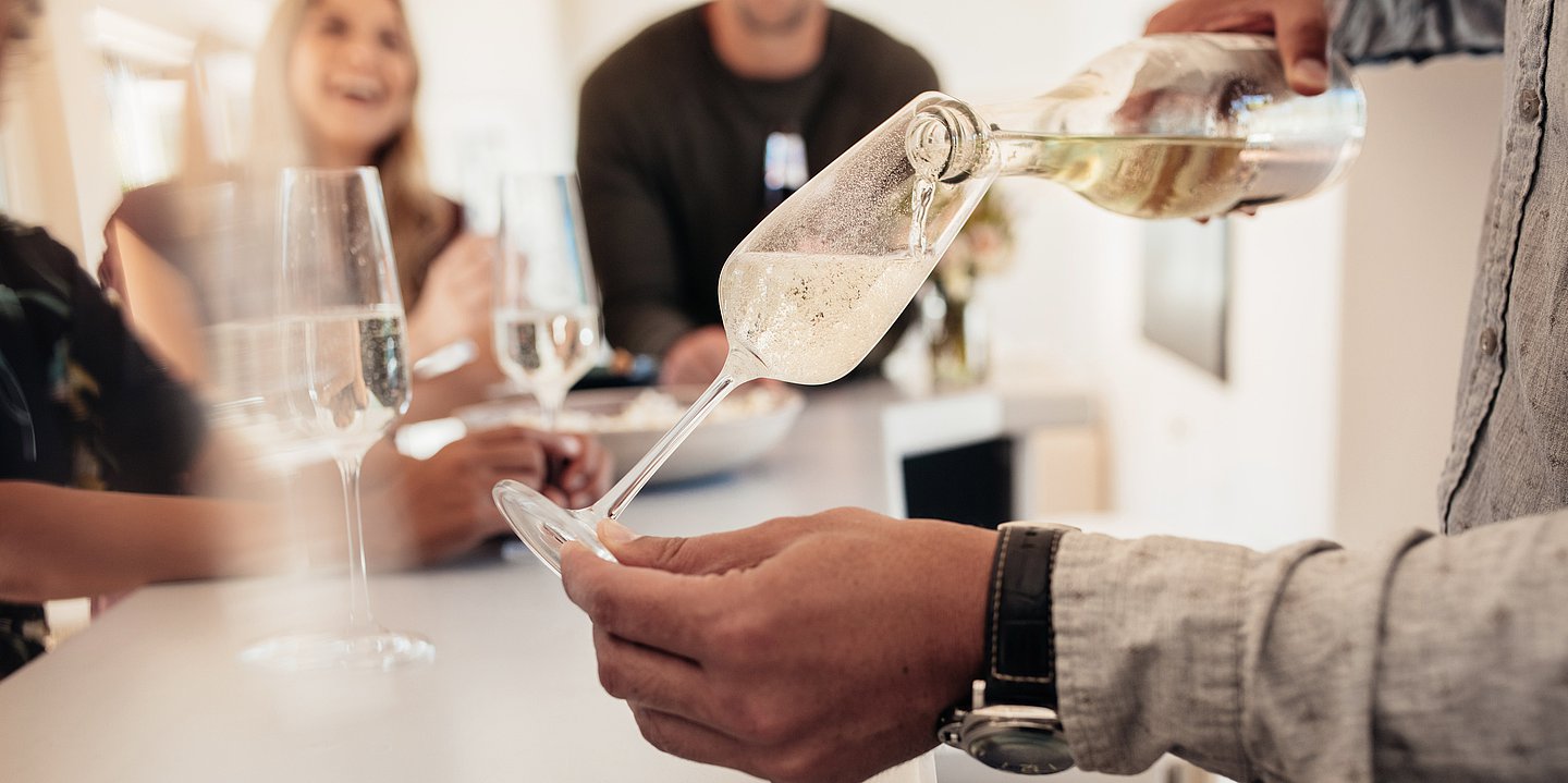 Auf einer Party stehen mehrere Personen im Kreis, alle haben Sektgläser in der Hand. Im Vordergrund steht ein Mann, der sich Sekt einschenkt.
