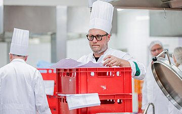 Koch mit wei&szlig;er M&uuml;tze arbeitet in einer Gro&szlig;k&uuml;che, weitere Mitarbeitende im Hintergrund.