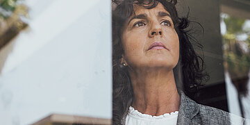 Foto: Eine Frau mit dunklen Haaren schaut aus dem Fenster. Das Foto ist von unten und linksb&uuml;ndig aufgenommen.
