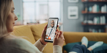 Eine Frau sitzt auf dem Sofa und h&auml;lt ein Handy vor sich. Auf dem Bildschirm ist ein Anruf mit einem Arzt zu sehen.