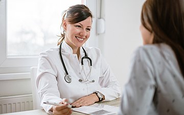 Eine Hausärztin sitzt an ihrem Praxistisch und ist im Gespräch mit einer Patientin, die ihr gegenüber sitzt.