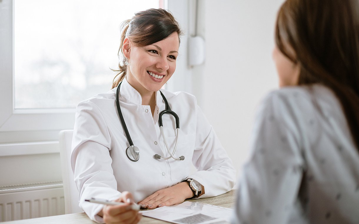 Eine Hausärztin sitzt an ihrem Praxistisch und ist im Gespräch mit einer Patientin, die ihr gegenüber sitzt.