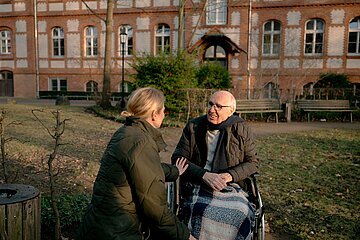 Eine Frau sitzt im Freien neben einem &auml;lteren Mann im Rollstuhl, der mit einer Decke bedeckt ist, und unterh&auml;lt sich mit ihm vor einem Geb&auml;ude.
