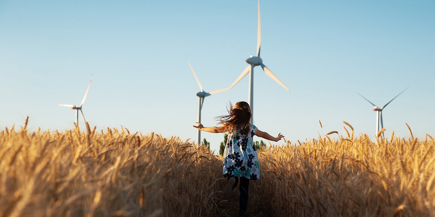 Ein Mädchen rennt durch ein Feld. In der Ferne stehen Windräder.