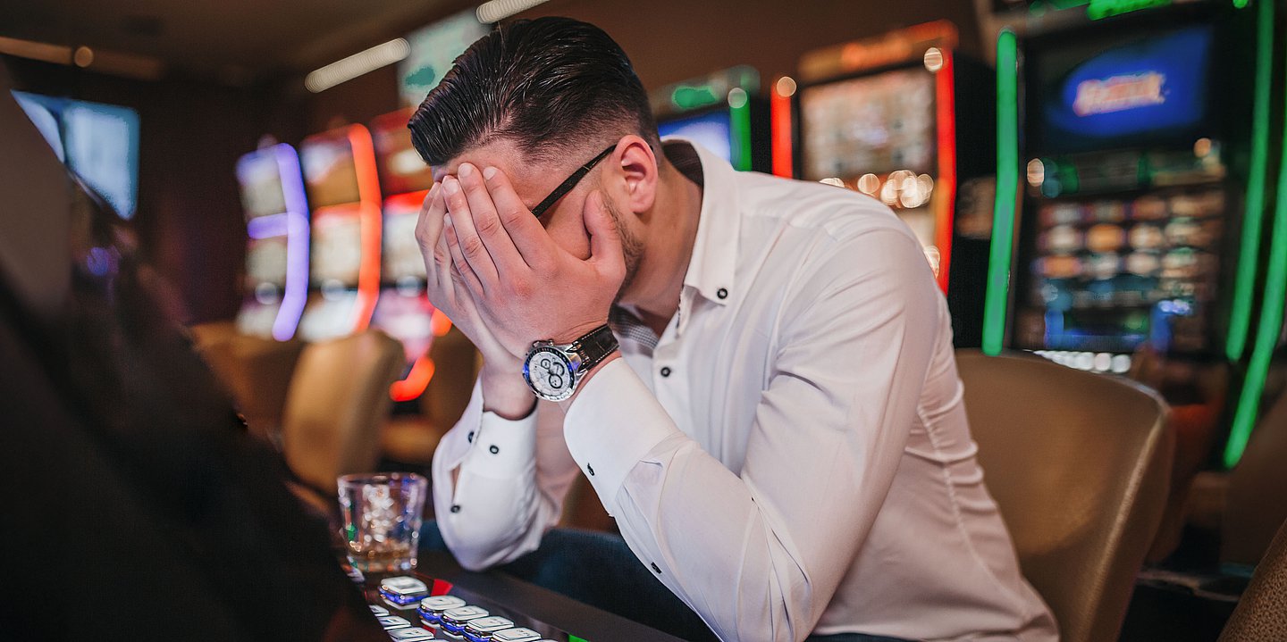 Mann vor Glücksspielautomat hält sich verzweifelt die Hände vors Gesicht.