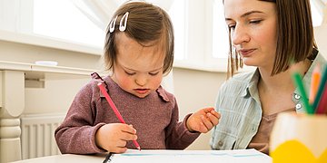 Alleinerziehende mit beeinträchtigten Kindern Foto einer jungen Mutter, die neben ihrem Kleinkind mit Trisomie 21 sitzt, das malt