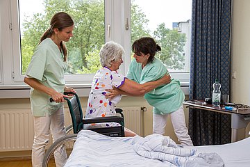 Zwei Pflegekräfte helfen einer älteren Frau sich in einen Rollstuhl zu setzten.