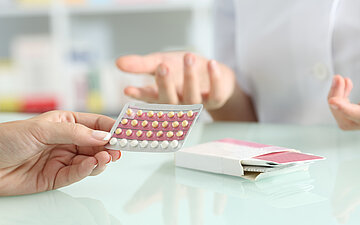 Foto zeigt einen Tisch auf dem eine Packung Antibabypillen liegt. Auf der Seite des Betrachters h&auml;lt eine Hand einen Blister mit den Pillen, auf der anderen Seite des Tisches sieht man den Ausschnitt einer Person in wei&szlig;em Kittel, deren H&auml;nde gestikulieren. Person 