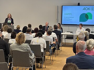 Das Bild zeigt eine Totale. Im Vordergrund mit dem R&uuml;cken zum Betrachter und zur Betrachterin die Teilnehmenden der Pressekonferenz. Im Hintergrund auf dem Podium die Protagonistinnen und Protagonisten: Dr. Martina Niemeyer, Clemens Hoch und Dr. Barabara R&ouml;mer
