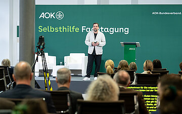 Der Moderator Michael Bernatek steht auf der B&uuml;hne und begr&uuml;&szlig;t das Publikum. 