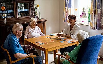An einem gro&szlig;en Holztisch sitzen drei Seniorinnen und Senioren mit einer Pflegerin. Sie spielen Karten.