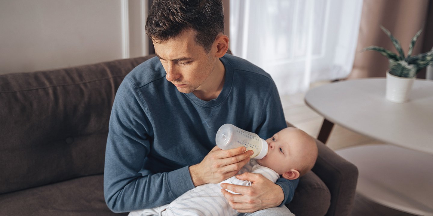Das Foto zeigt einen Mann mit grauem Pullover, der auf dem Sofa ein Baby mit der Flasche stillt, das in seinem Schoß liegt. Der Mann schaut traurig zur Seite und blickt nicht auf das Kind.