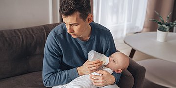 Postpartale Depression bei Männern Das Foto zeigt einen Mann mit grauem Pullover, der auf dem Sofa ein Baby mit der Flasche stillt, das in seinem Schoß liegt. Der Mann schaut traurig zur Seite und blickt nicht auf das Kind.
