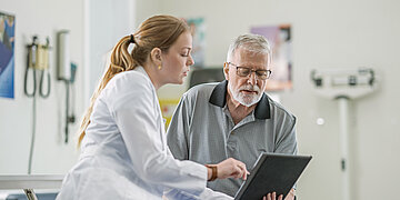 Eine &Auml;rztin zeigt einem &auml;lteren Patienten in einem Behandlungsraum Informationen auf einem Tablet. Beide blicken konzentriert auf den Bildschirm.