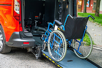 Die offene Heckklappe eines roten Wagens ist sichtbar, hier ist eine mobile Rollstuhlrampe befestigt, auf der ein leerer Rollstuhl steht, um in den Wagen gehoben zu werden.