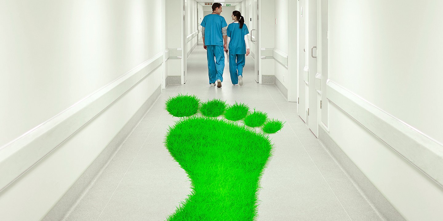 Foto: Ein Krankehausflur mit zwei Pflegenden von hinten und einem großen grünen Fußabdruck auf dem Fußboden.