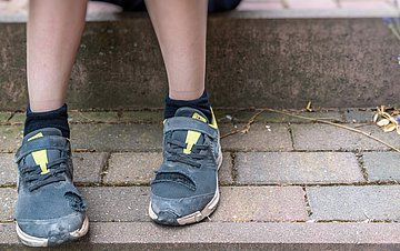 Kinderarmut Foto, auf dem die nackten Beine und abgenutzten Schuhe eines Kindes zu sehen sind, das auf einer steinernen Stufe sitzt