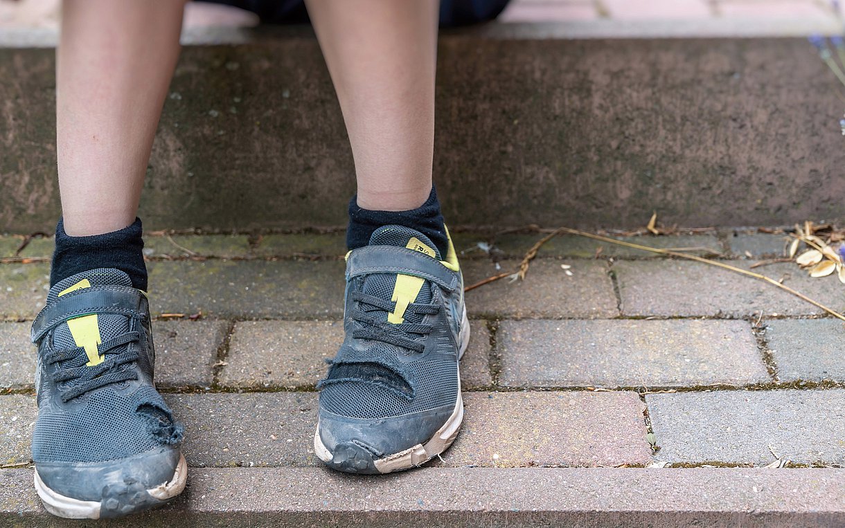 Foto, auf dem die nackten Beine und abgenutzten Schuhe eines Kindes zu sehen sind, das auf einer steinernen Stufe sitzt