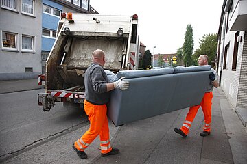 Das Foto zeigt zwei M&uuml;llweker in klassischer orangefarbener Arbeitskleidung. Sie tragen ein ausgedientes hellblaues Sofa zum M&uuml;llwagen, der im Hintergrund steht.