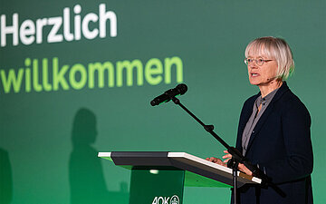 Eine Frau h&auml;lt eine Rede hinter einem Rednerpult. 