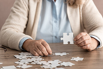 Eine am Tische sitzende Person h&auml;lt ein Puzzle-Teil in der linken Hand etwas &uuml;ber den Tisch hoch. Mit der rechten Hand verharrt auf dem Tisch, auf dem weitere Puzzleteile liegen, um das n&auml;chste Teil zu nehmen.
