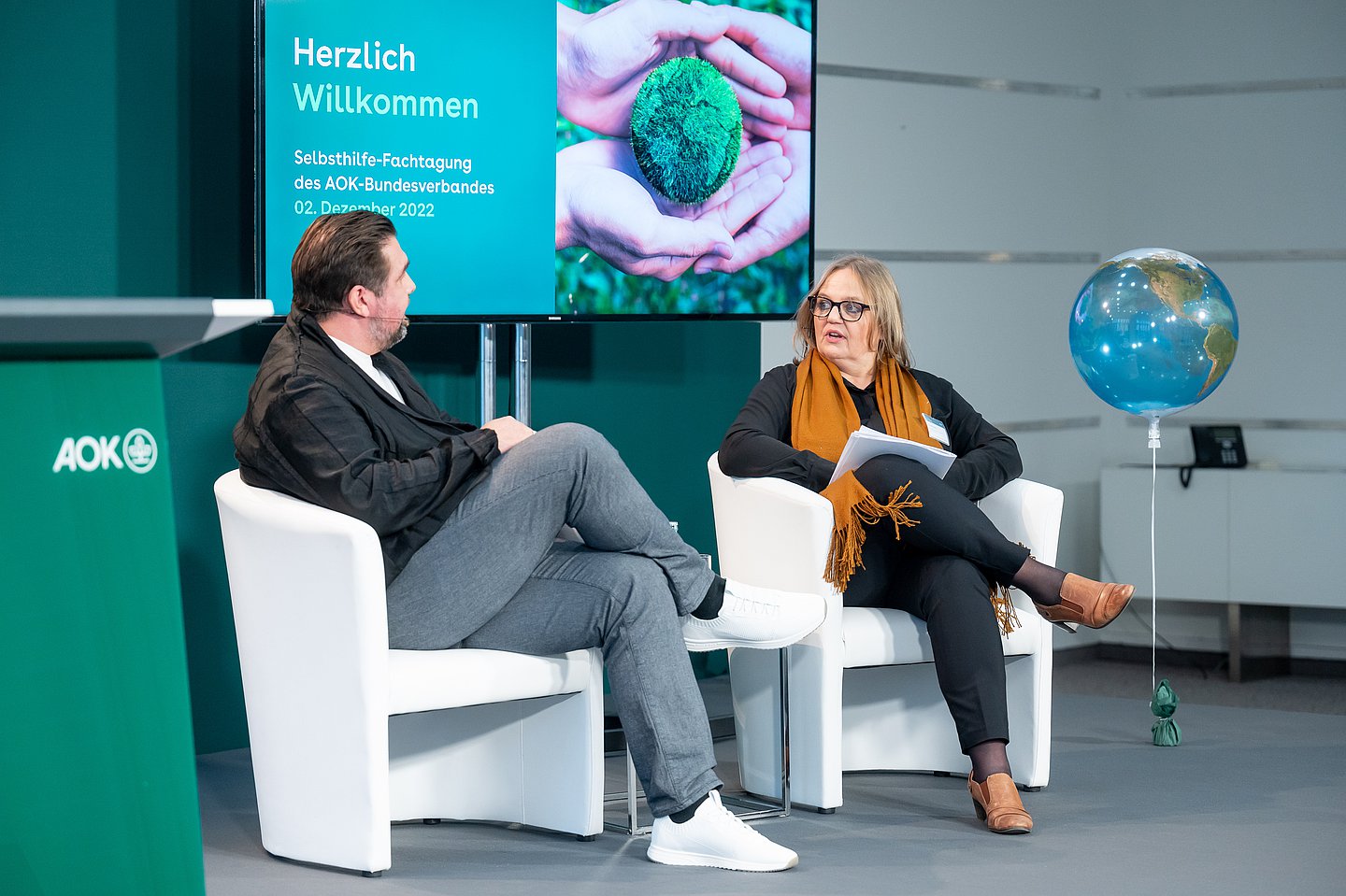 Moderator Michael Bernatek und Claudia Schick, Selbsthilfereferentin des AOK-Bundesverbandes, sitzen in weißen Sesseln auf dem Podium der Fachtagung.