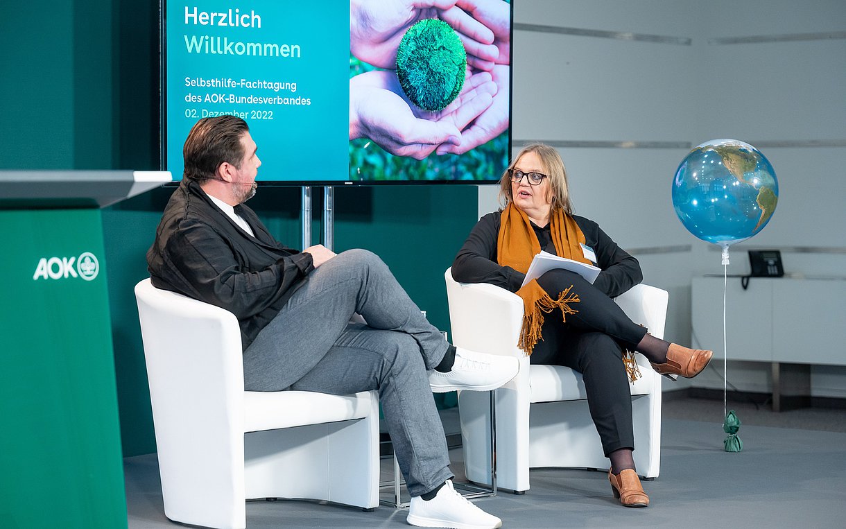 Moderator Michael Bernatek und Claudia Schick, Selbsthilfereferentin des AOK-Bundesverbandes, sitzen in weißen Sesseln auf dem Podium der Fachtagung.