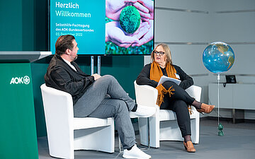 Moderator Michael Bernatek und Claudia Schick, Selbsthilfereferentin des AOK-Bundesverbandes, sitzen in wei&szlig;en Sesseln auf dem Podium der Fachtagung.