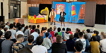 Das Foto zeigt eine Theaterauff&uuml;hrung in einer Turnhalle. Im Hintergrund sieht man die B&uuml;hne, im Vordergrund eine Gruppe von Sch&uuml;lerinnen und Sch&uuml;lern, die gespannt zuschauen.