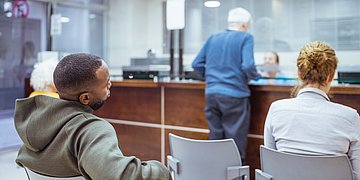 Auf Stühlen vor einem Tresen sitzen drei Personen: Ein Mann, eine Frau und eine ältere Dame. Am Tresen steht ein älterer Herr und spricht mit der Sprechstundenhilfe hinter dem Tresen.