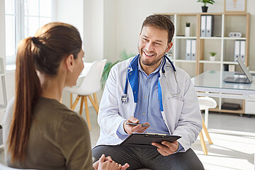 Eine Patientin im Sprechzimmer des Arztes. Beide sitzen sich gegen&uuml;ber und sind im Gespr&auml;ch miteinander, der Arzt h&auml;lt dabei ein Klemmbrett in der einen und eine Schreiber in der anderen Hand.