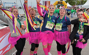 Im Vordergrund steht ein jubelndes Frauenteam in einem farbenfrohen Outfit. Im Hintergrund ist links die F&ouml;rde zu sehen, mittig der Zielbogen des Laufs mit ankommenden L&auml;ufern und L&auml;uferinnen sowie am rechten Seitenrand die r&uuml;ckw&auml;rtige Ansicht der wei&szlig;en Pagodenzelte der Teams.