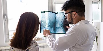 Osteoporos Röntgenbild Wirbelsäule Das Foto zeigt einen Arzt mit Brille, Bart und dunklen Haaren, der vor einem Fenster einer brünetten Patientin mit Zopf ein Röntgenbild einer Wirbelsäule erläutert.